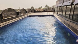 A swimming pool with clear blue water, surrounded by a fence and a building with large windows. - Hotel Lotus Paradise (Sonepat)