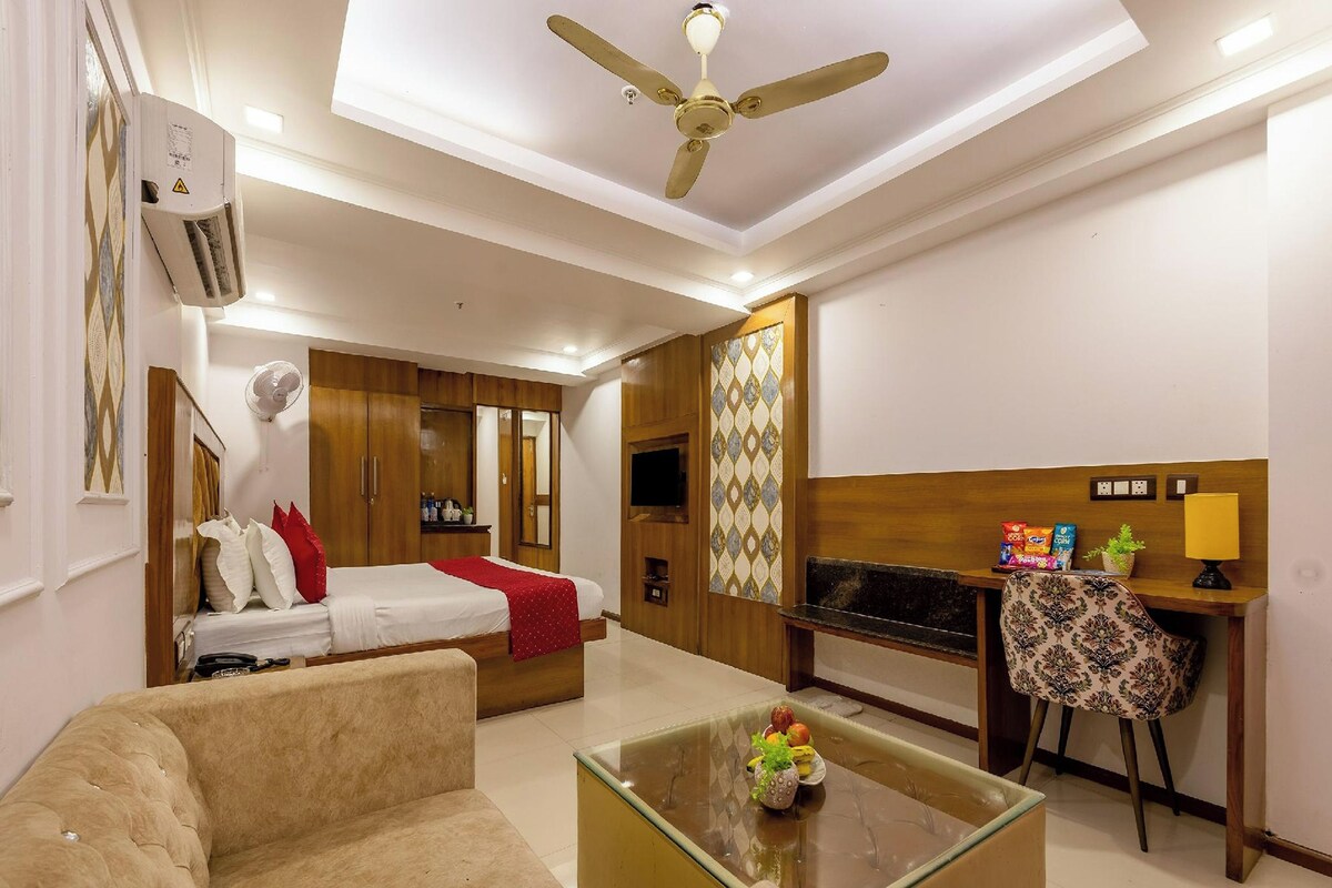 A hotel room with a bed, a sofa, a glass coffee table, a desk with a chair, a ceiling fan, and a TV.