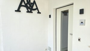 A hallway with a white wall, a door with a glass panel, and a black A&J logo on the wall. - A&A Rooms (Anacapri)