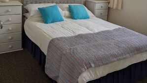 Une chambre bien rangée, avec un grand lit, deux tables de chevet, une commode, un tableau encadré au mur et une fenêtre avec des rideaux.