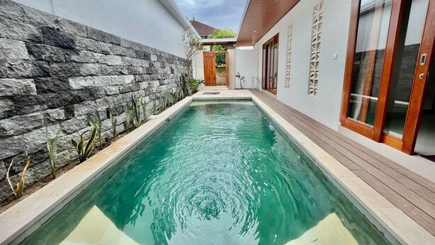 A swimming pool with a stone wall on one side and wooden deck on the other, leading to a wooden door. - Belvilla Tirta Sari Resort Central Ubud (Ubud)