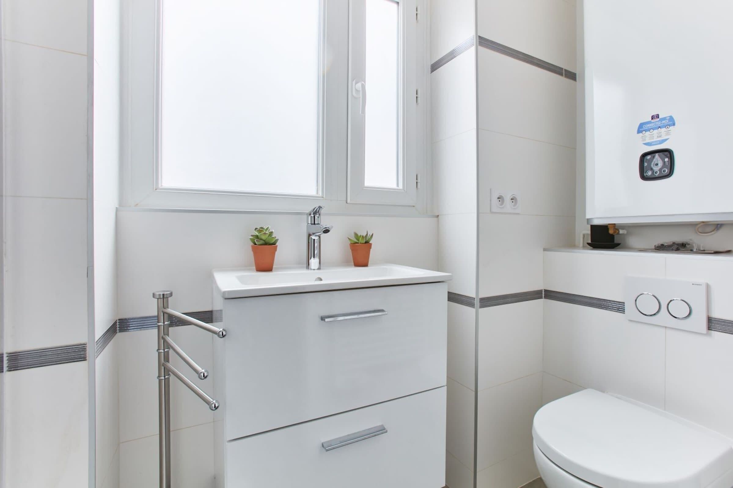 Une salle de bain moderne avec un lavabo blanc, deux plantes en pot, des toilettes et un chauffe-eau.