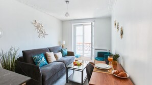 A modern living room with a sofa, coffee table, and dining table. There is a balcony with a view, a plant, and a lamp.