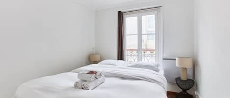 A neatly made bed with white linens and rolled towels, a window with a view of a balcony, and two bedside lamps on a nightstand.