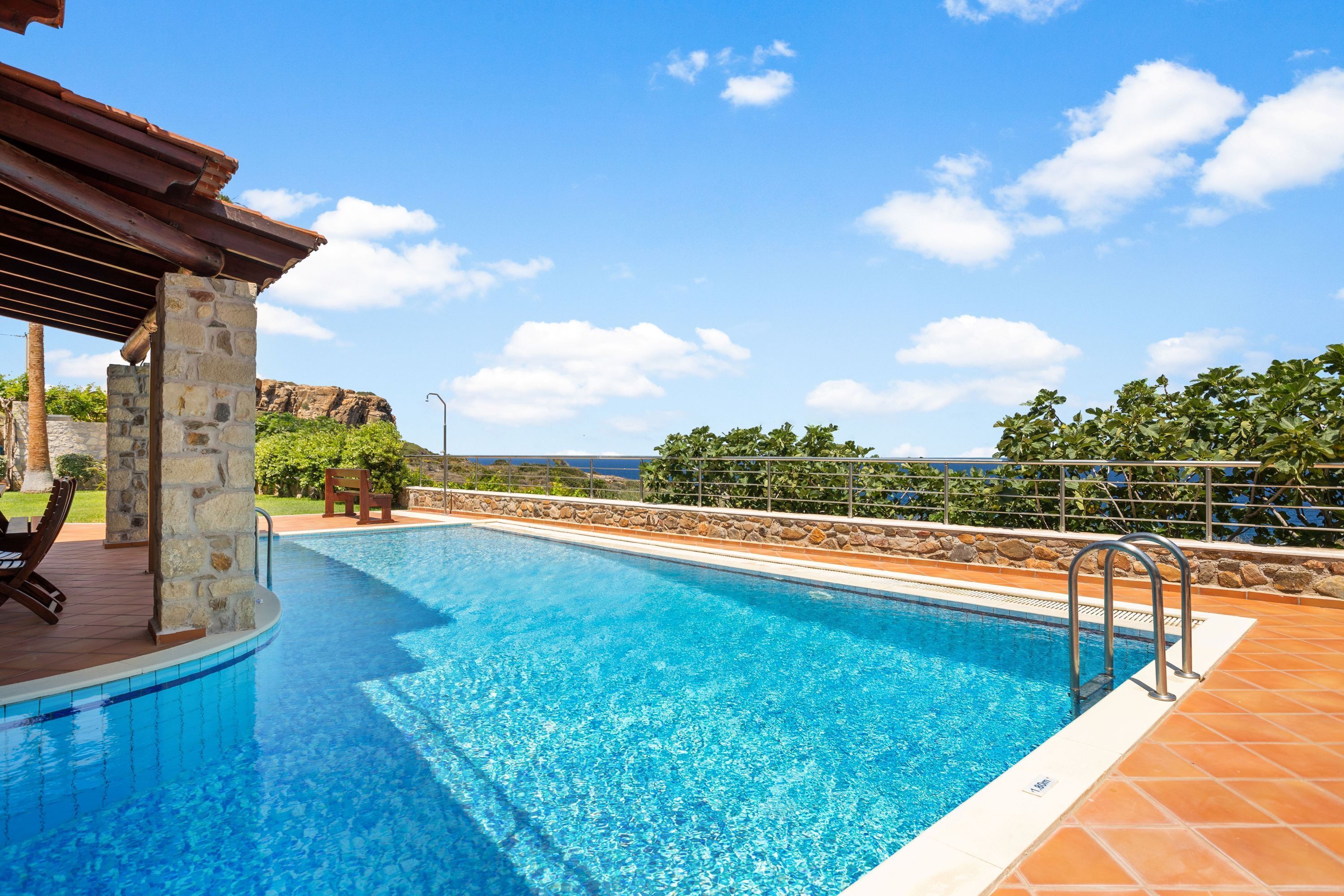 A swimming pool with a stone wall and a wooden deck, surrounded by a stone wall and a view of the ocean.