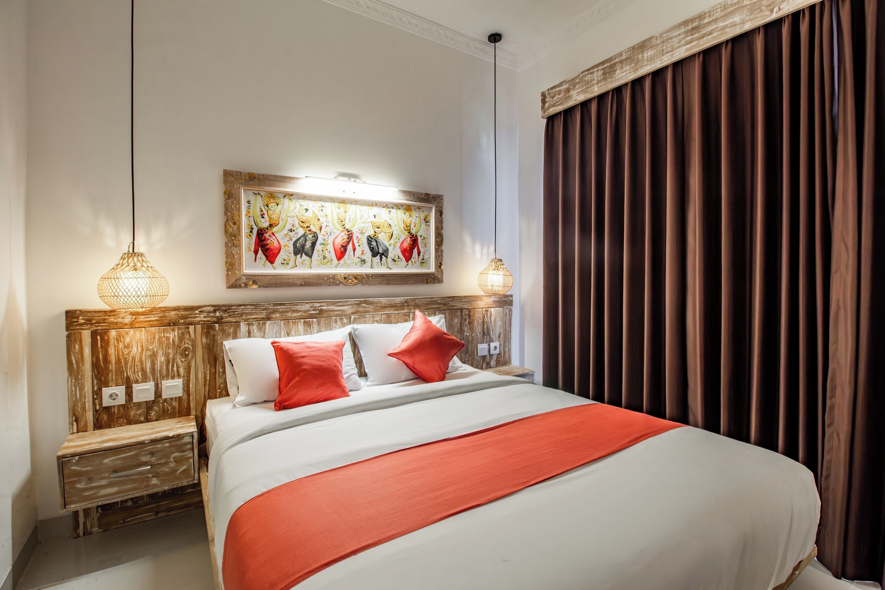 A hotel room with a wooden headboard, a large bed with red and white bedding, two bedside lamps, a framed artwork above the bed, and a window with curtains.