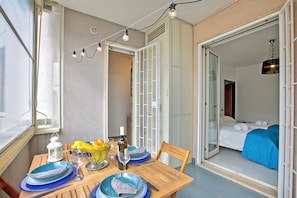Un coin repas avec une table dressée pour deux personnes, une fenêtre et une vue sur une chambre à coucher avec un lit et une couverture bleue.