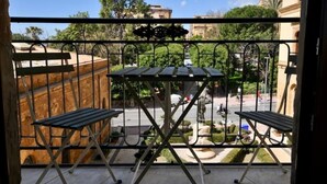 Un balcon doté de deux tables et de deux chaises en bois, d’une balustrade en fer forgé et offrant une vue sur une rue bordée d’immeubles et de verdure.