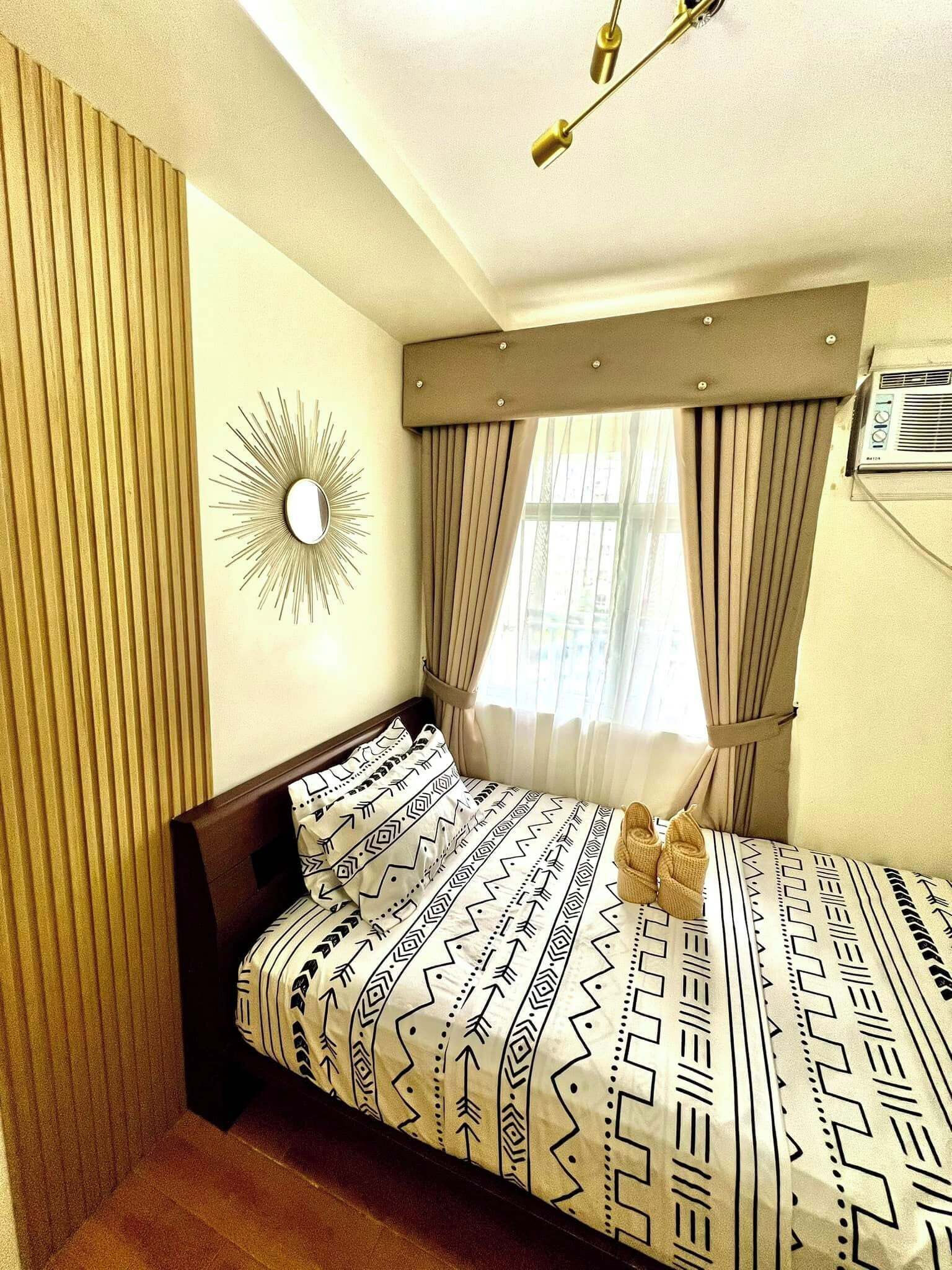 A hotel room with a bed, a wall-mounted mirror, a window with curtains, and an air conditioning unit.