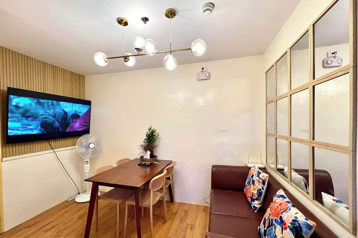 Una sala moderna con piso de madera, un sofá de cuero marrón con cojines estampados, una mesa de comedor de madera con sillas, un televisor de pantalla plana montado en la pared, una lámpara en el techo y una ventana con persianas.