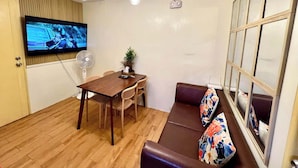 A compact living space with a wooden floor, a dining table with chairs, a sofa with floral cushions, a TV mounted on the wall, and a ceiling light fixture. - Seawind Condo 2 BR Near Airport & Samal (Davao)