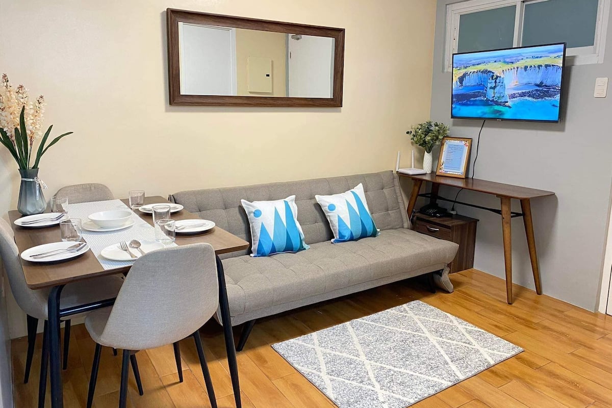 Una sala de estar moderna con un sofá, una mesa de comedor y sillas. Hay un televisor de pantalla plana montado en la pared, un escritorio pequeño con una silla, y un jarrón decorativo con flores.