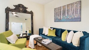 A living room with a large ornate mirror, a blue sofa with multiple cushions, a green armchair, and a painting of a forest scene on the wall. - Cozy 3 bedroom - Free Parking, close to buses.  (Boston)