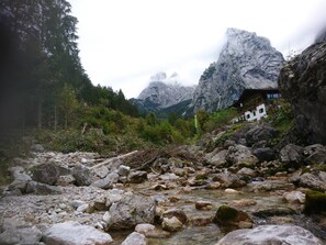 Un chalet de montagne rustique, avec un ruisseau rocheux en contrebas, entouré d’une forêt dense et de montagnes imposantes.