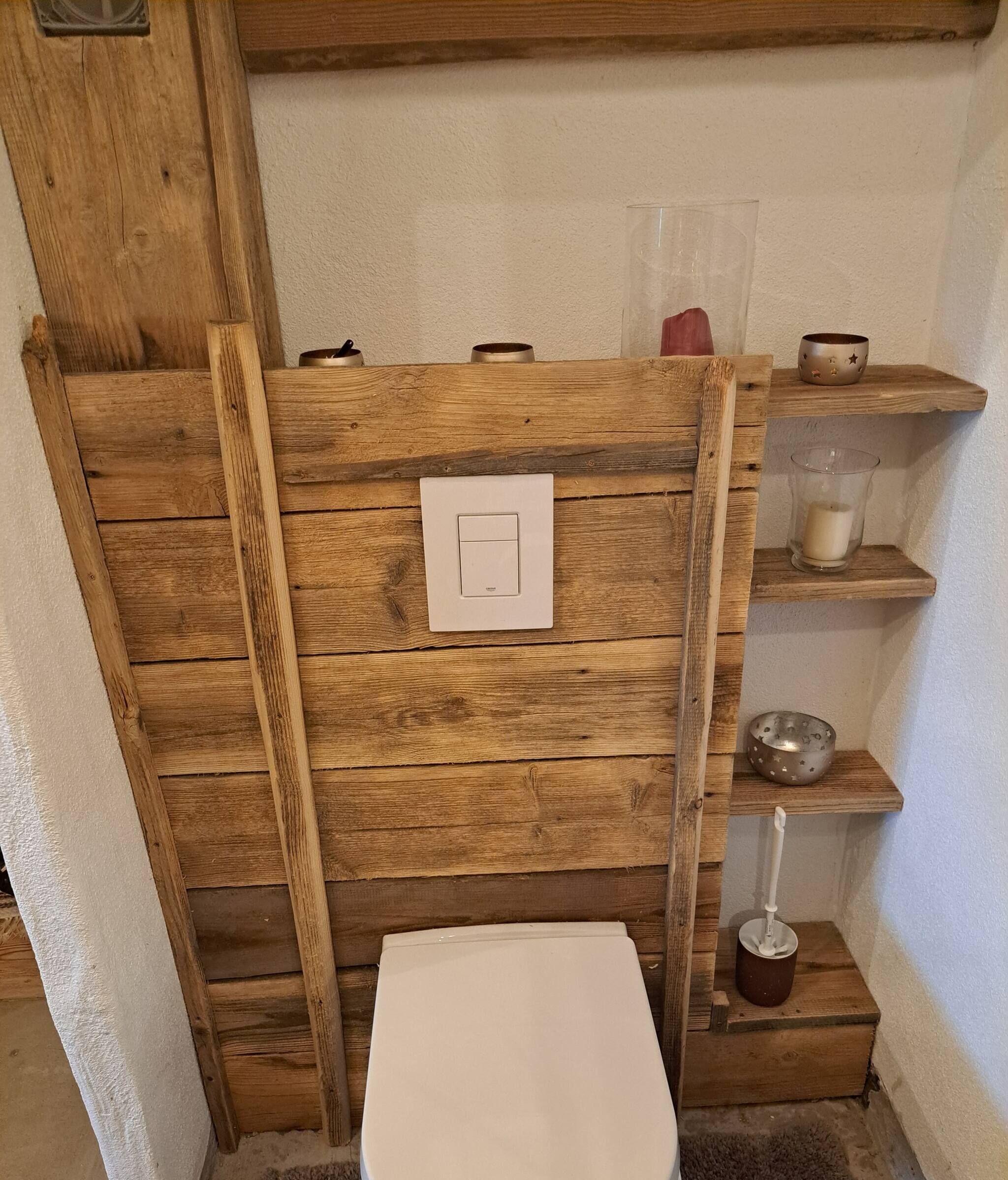 Ein rustikales Badezimmer mit einem Holzlagerfläschchen, einer weißen Toilette und Ablagen mit Pflegeprodukten.