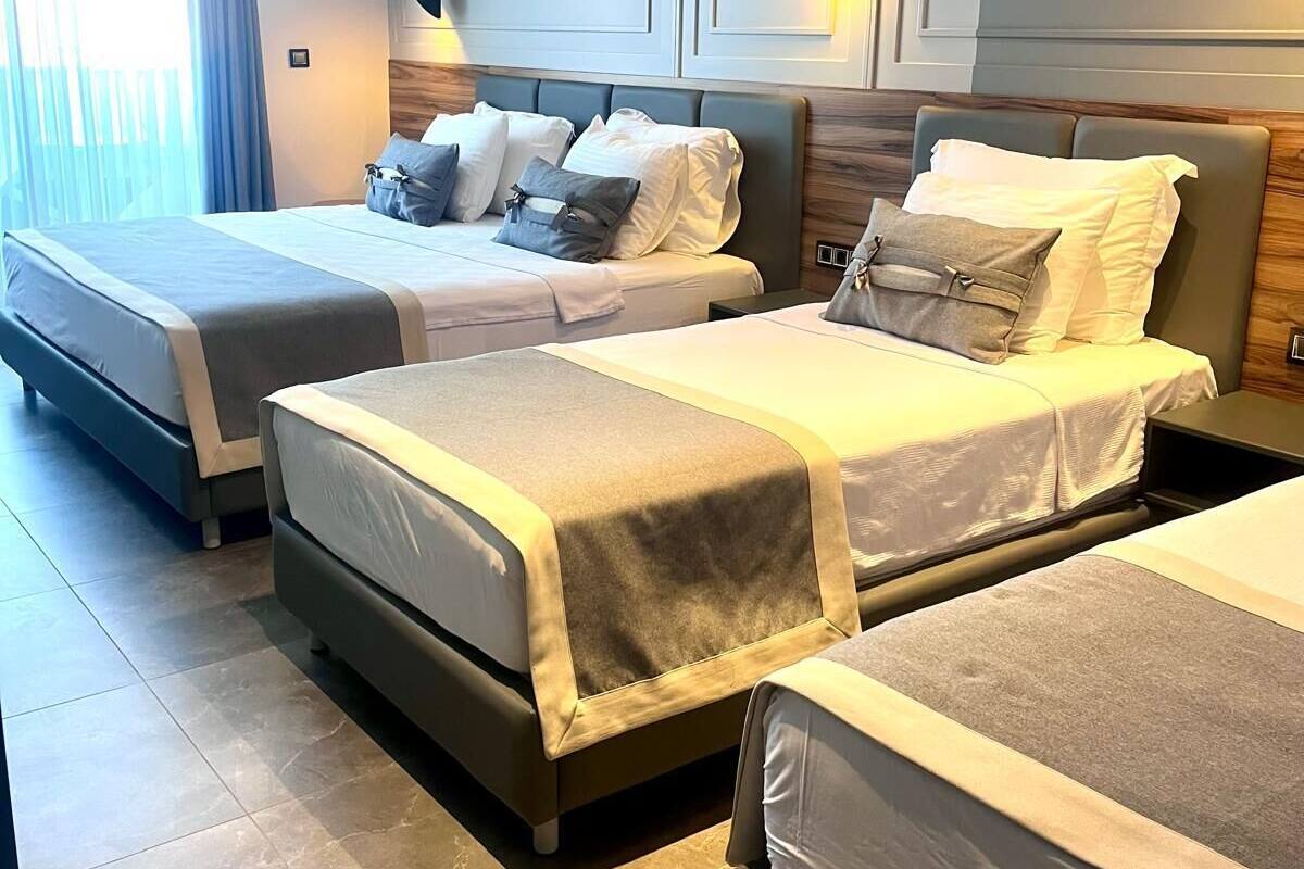A hotel room with three beds arranged in a row, a large window with curtains, and a modern light fixture.