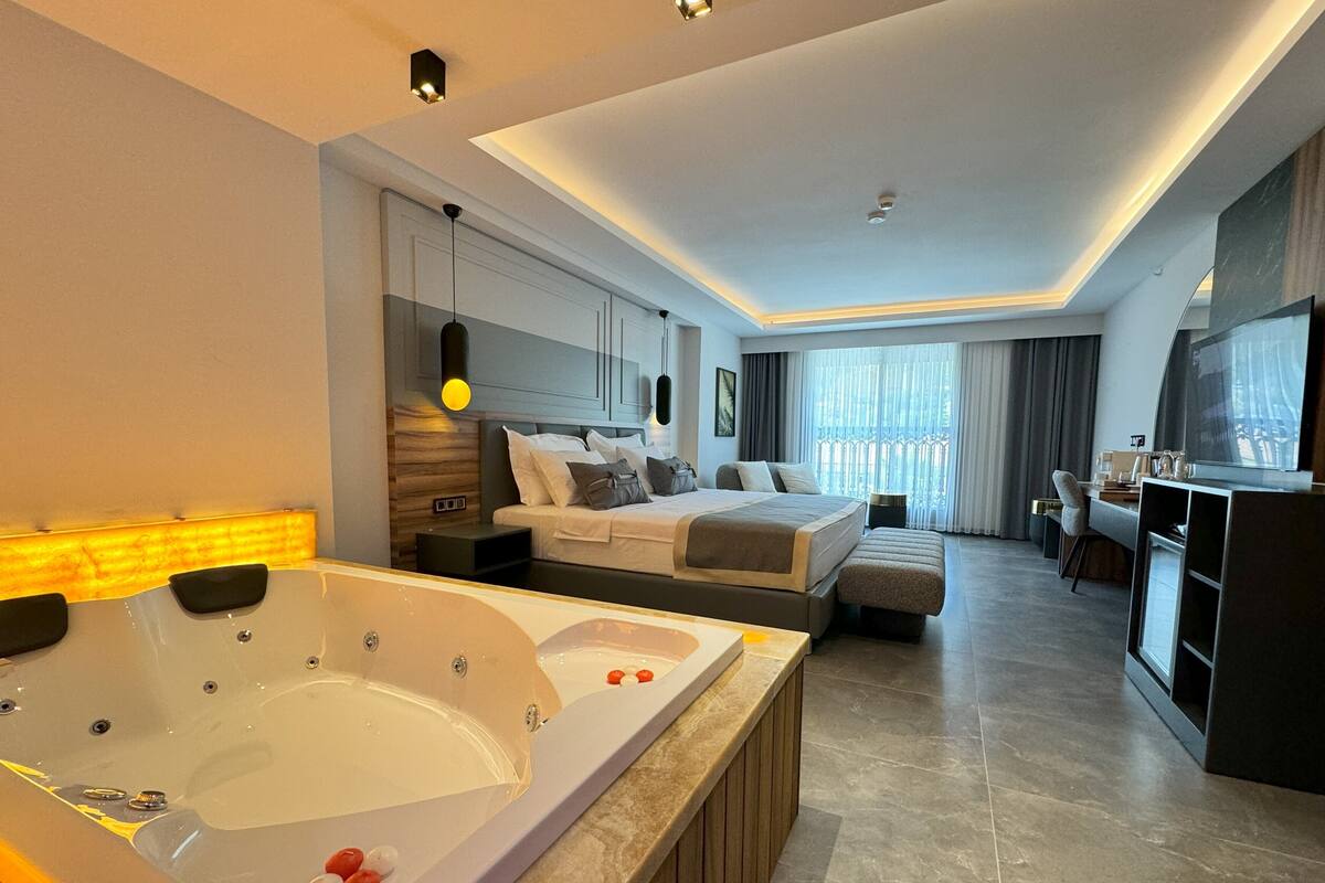A modern hotel room with a large bathtub, a bed, a desk, and a television.