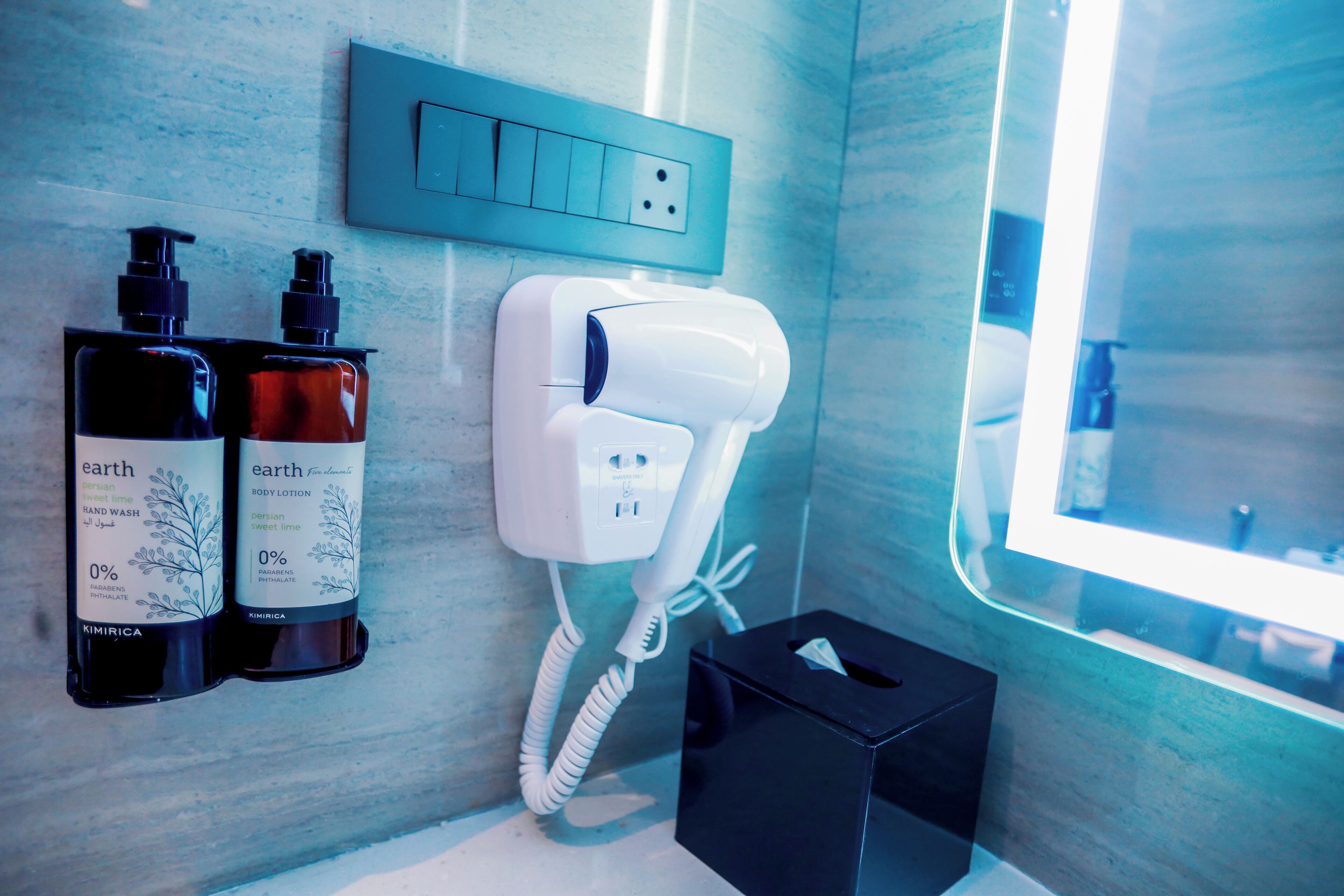 A bathroom with a hand dryer, two bottles of body lotion, and a trash bin.