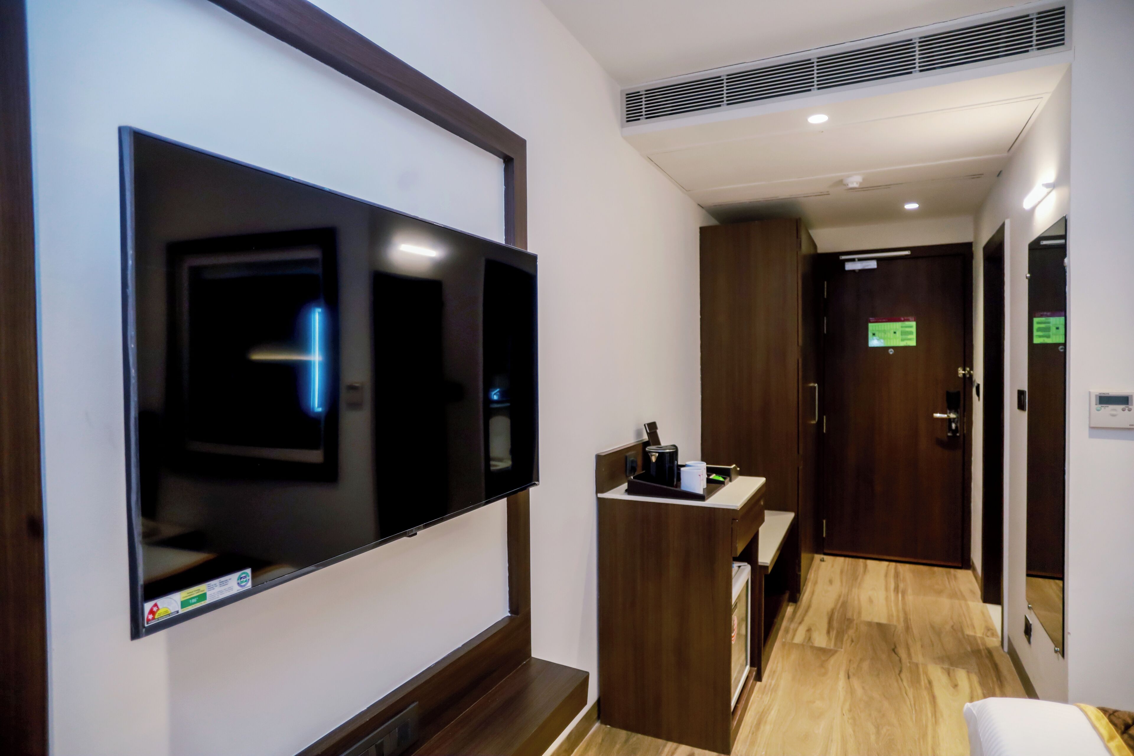 Passadís d'hotel amb un televisor de pantalla plana penjat a la paret, un taulell de recepció de fusta i una porta amb un rètol verd.