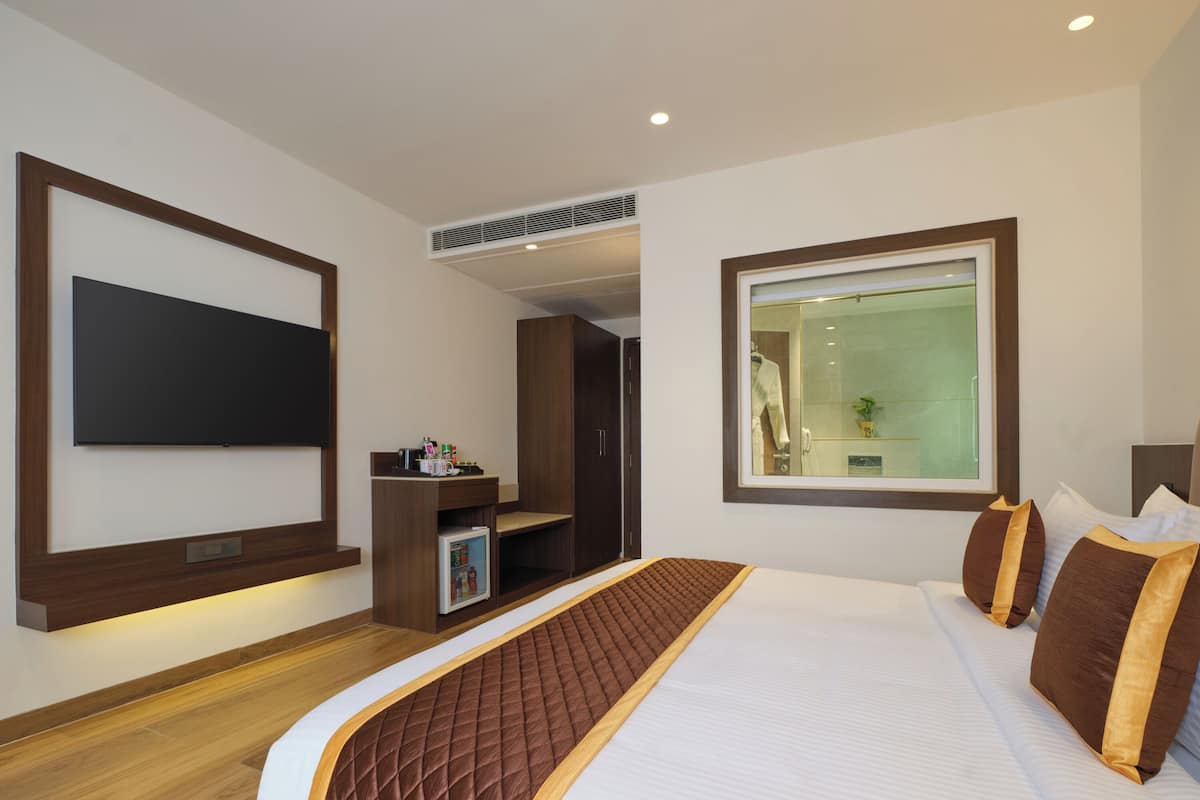 A hotel room with a large bed, a flat-screen TV mounted on the wall, a small kitchenette, and a large window with a view of the bathroom.
