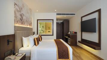 Quarto de hotel com uma cama grande, televisão presa à parede, secretária com telefone e banheiro visível através de uma porta de vidro.
