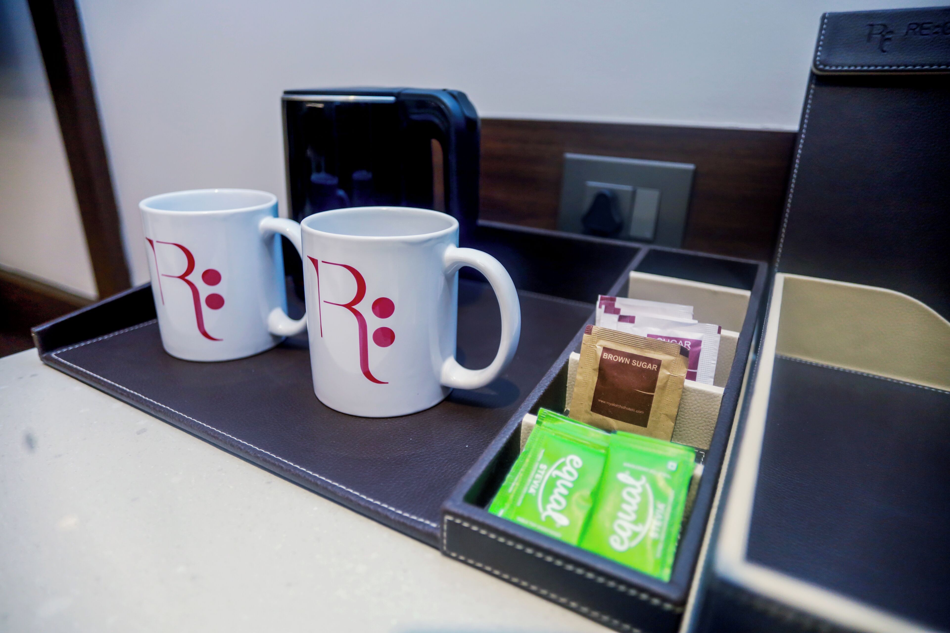 Una bandeja con dos tazas blancas con texto rojo, un teléfono inteligente, y una caja que contiene sobres verdes y un sobre de azúcar moreno.
