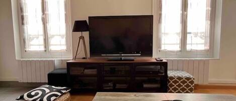 A living room with a TV on a wooden stand, a lamp, a tray with a cup, and a patterned cushion on the floor.