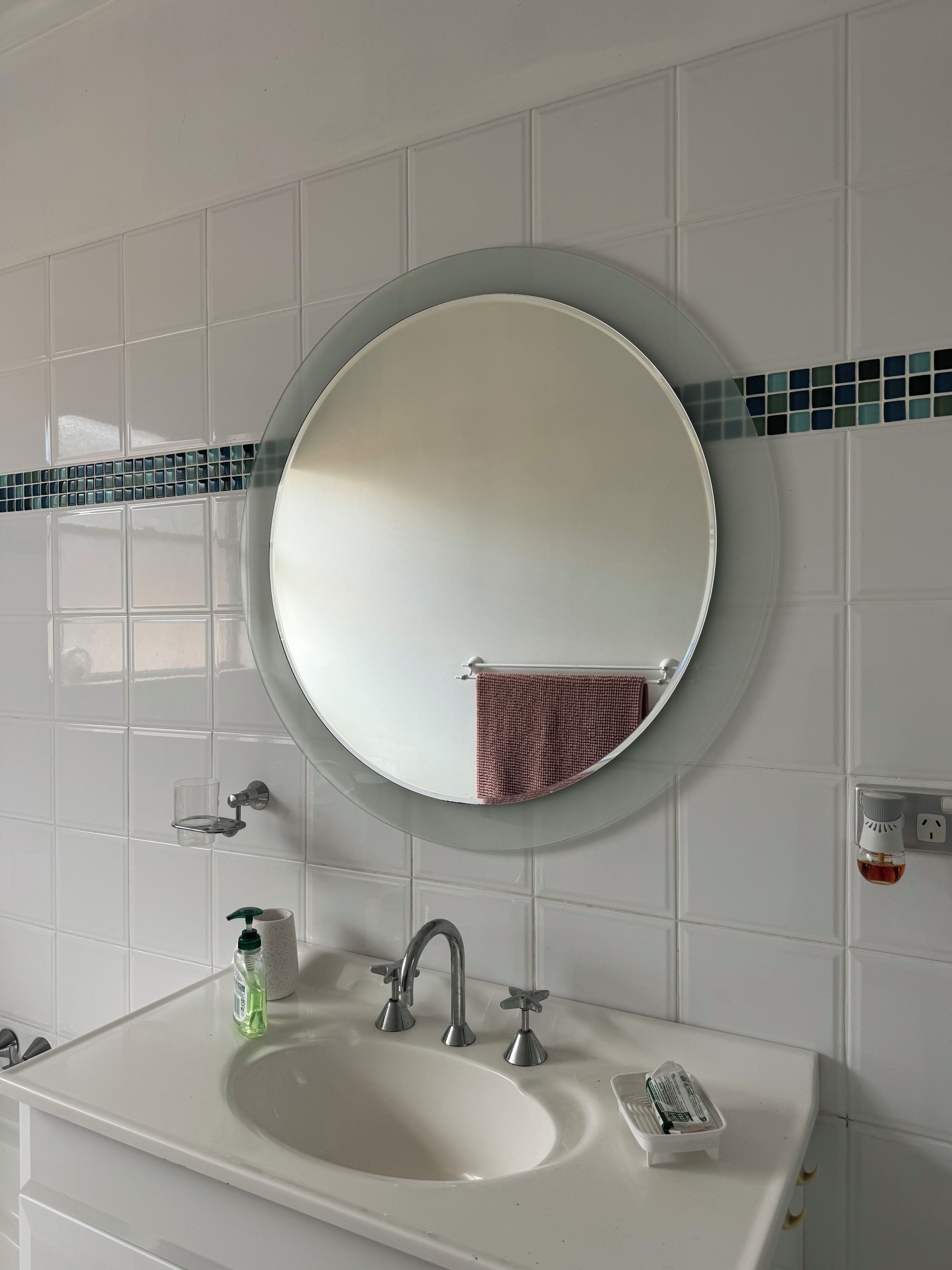 A bathroom with a white sink, a round mirror, a towel hanging on the wall, and a soap dispenser.