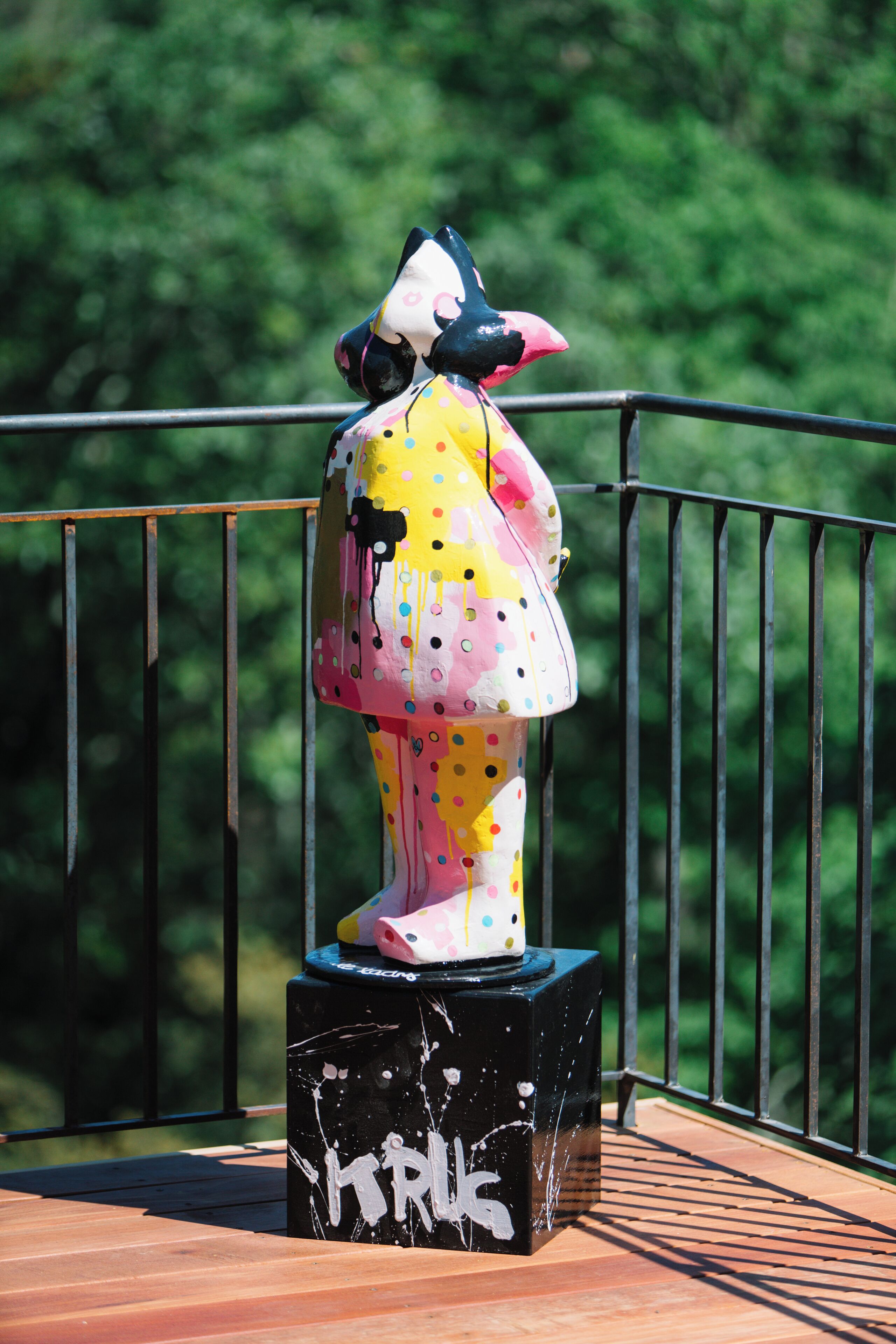 Une sculpture aux couleurs vives, posée sur un piédestal noir orné de graffitis, se dresse devant une rambarde métallique, le tout sur un fond vert flou.