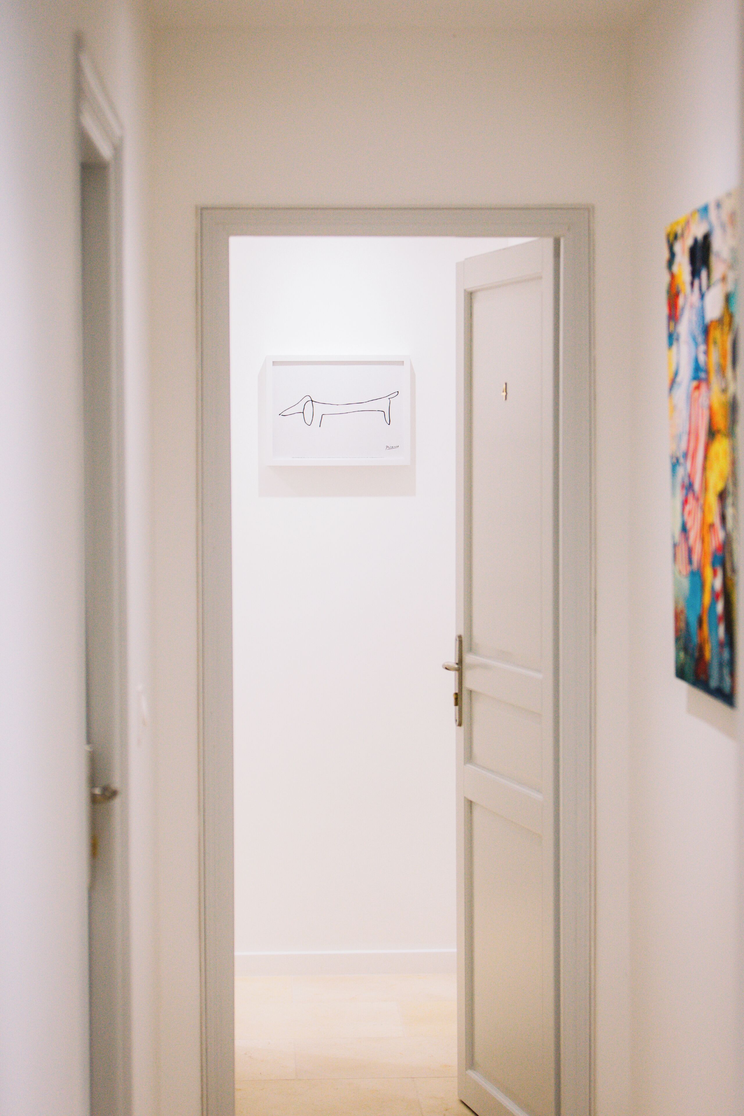 Un couloir avec une porte donnant sur une pièce, un tableau encadré au mur et une œuvre d’art colorée sur le côté droit.