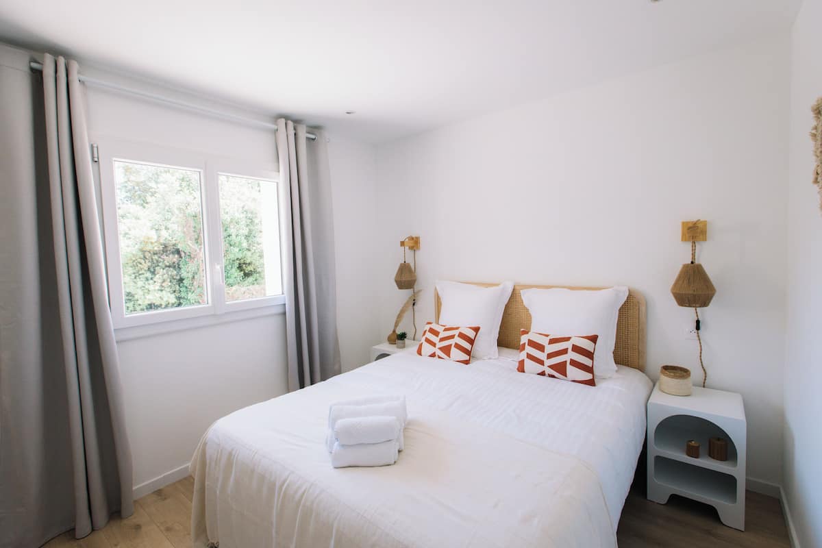 A neatly made bed with white linens and decorative pillows, a bedside table with a lamp, and a window with curtains.