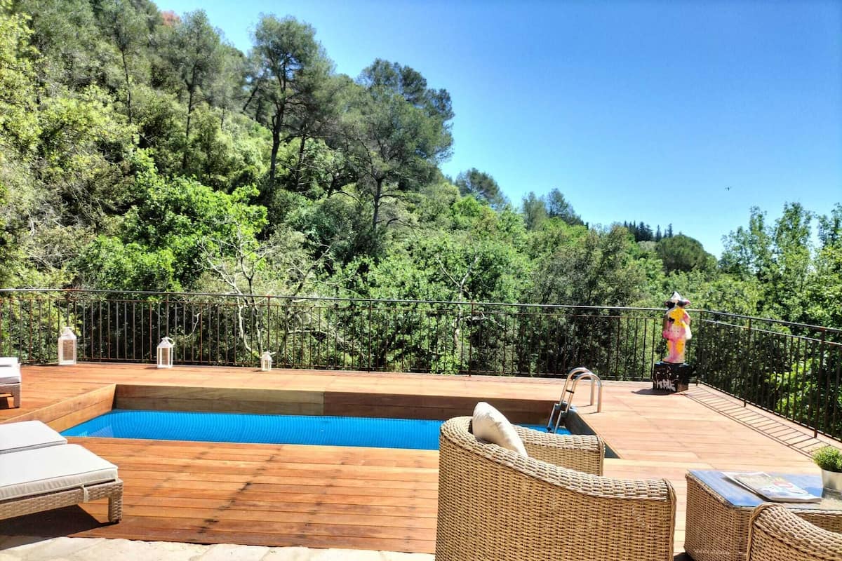A wooden deck with a swimming pool, wicker furniture, and a view of a forested hillside.