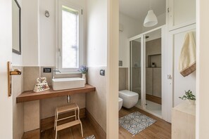 A compact bathroom with a wooden vanity, a small stool, a white sink, and a shower area.
