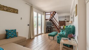 Una sala de estar con una escalera de madera, un sofá, un sillón verde y una mesita con un libro y flores.