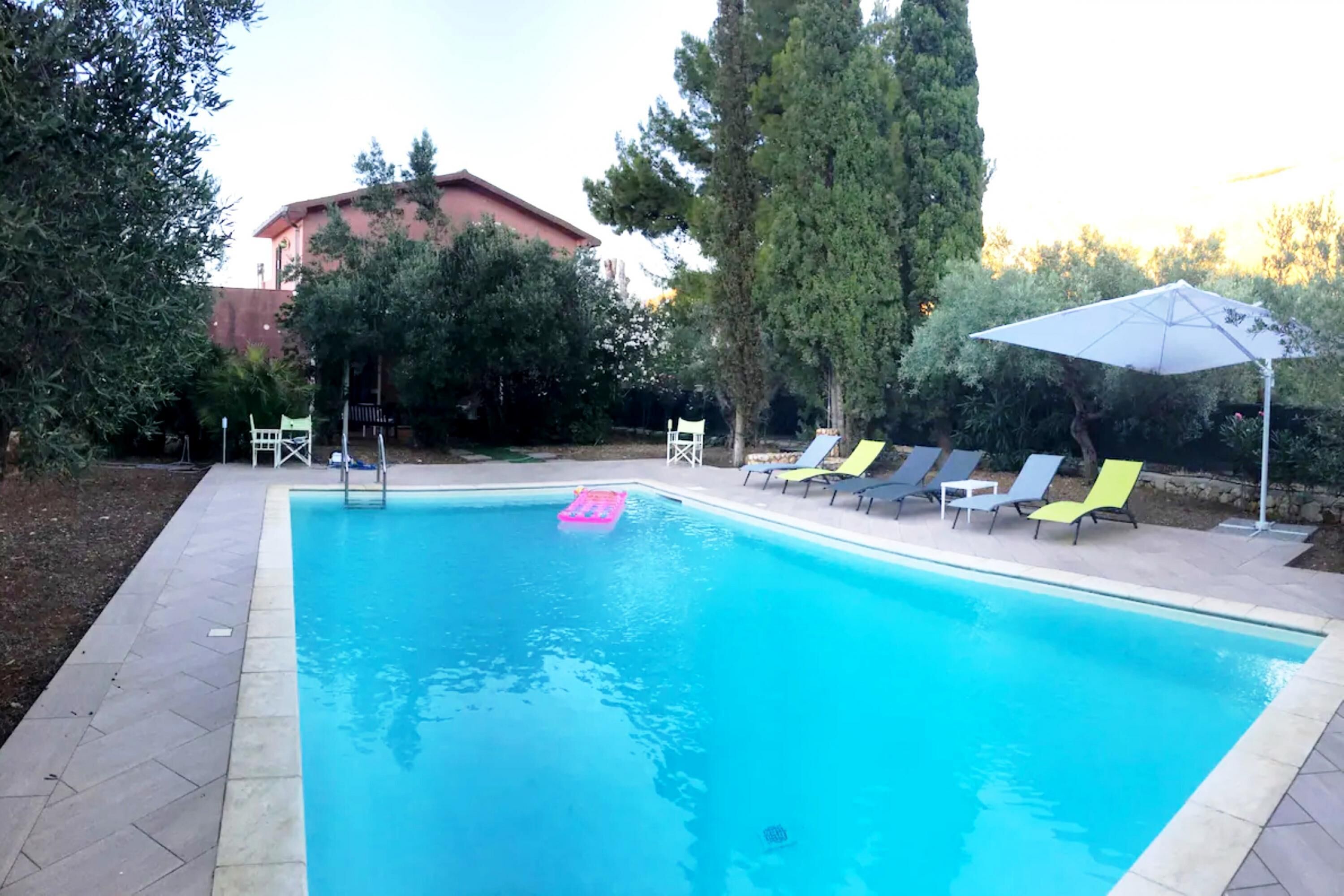 A rectangular swimming pool with a tiled edge, surrounded by a paved deck. There are several lounge chairs with umbrellas, and a tall tree is visible to the right.