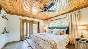 A bedroom with a wooden ceiling, a bed with a blue headboard, a bench, a nightstand with a lamp, and a window with curtains. - Broken Bow Cabin ~ 5 Mi to State Park w/ Hot Tub! (Broken Bow)