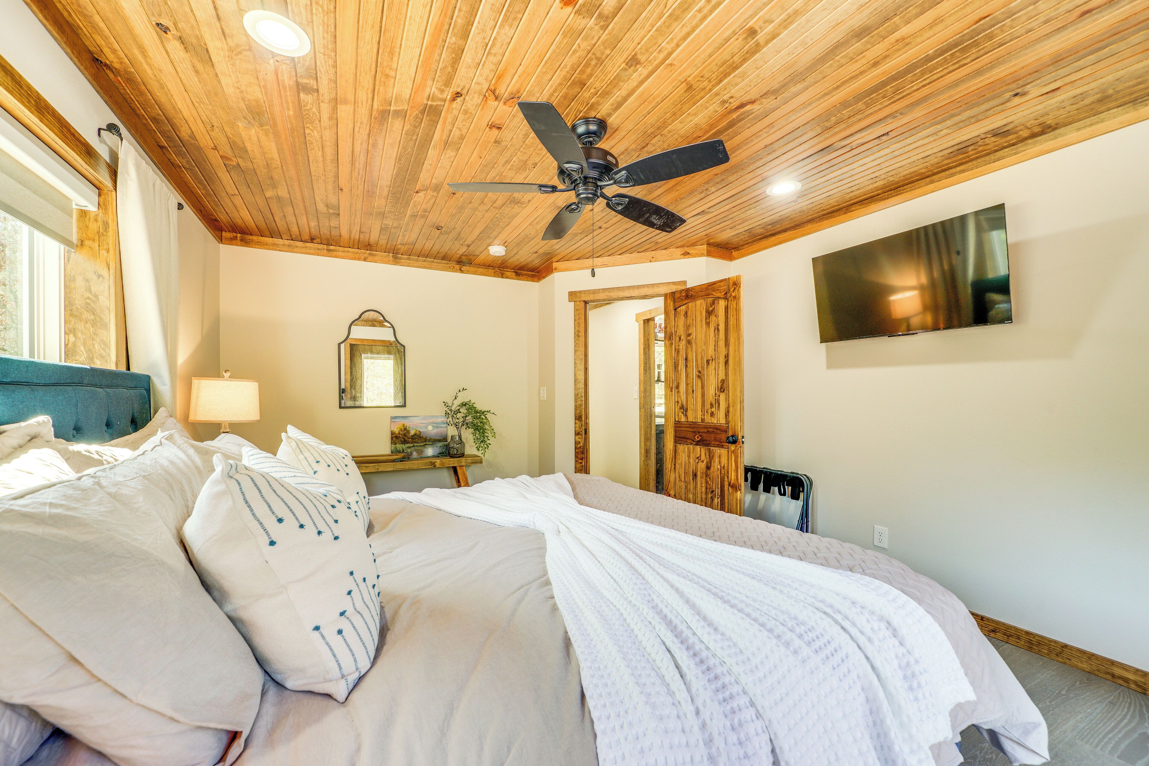 A bedroom with a wooden ceiling, a bed with white bedding, a ceiling fan, a flat-screen TV, and a wooden door.