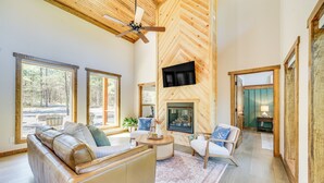 A spacious living room with a fireplace, large windows, and a ceiling fan. - Broken Bow Cabin ~ 5 Mi to State Park w/ Hot Tub! (Broken Bow)