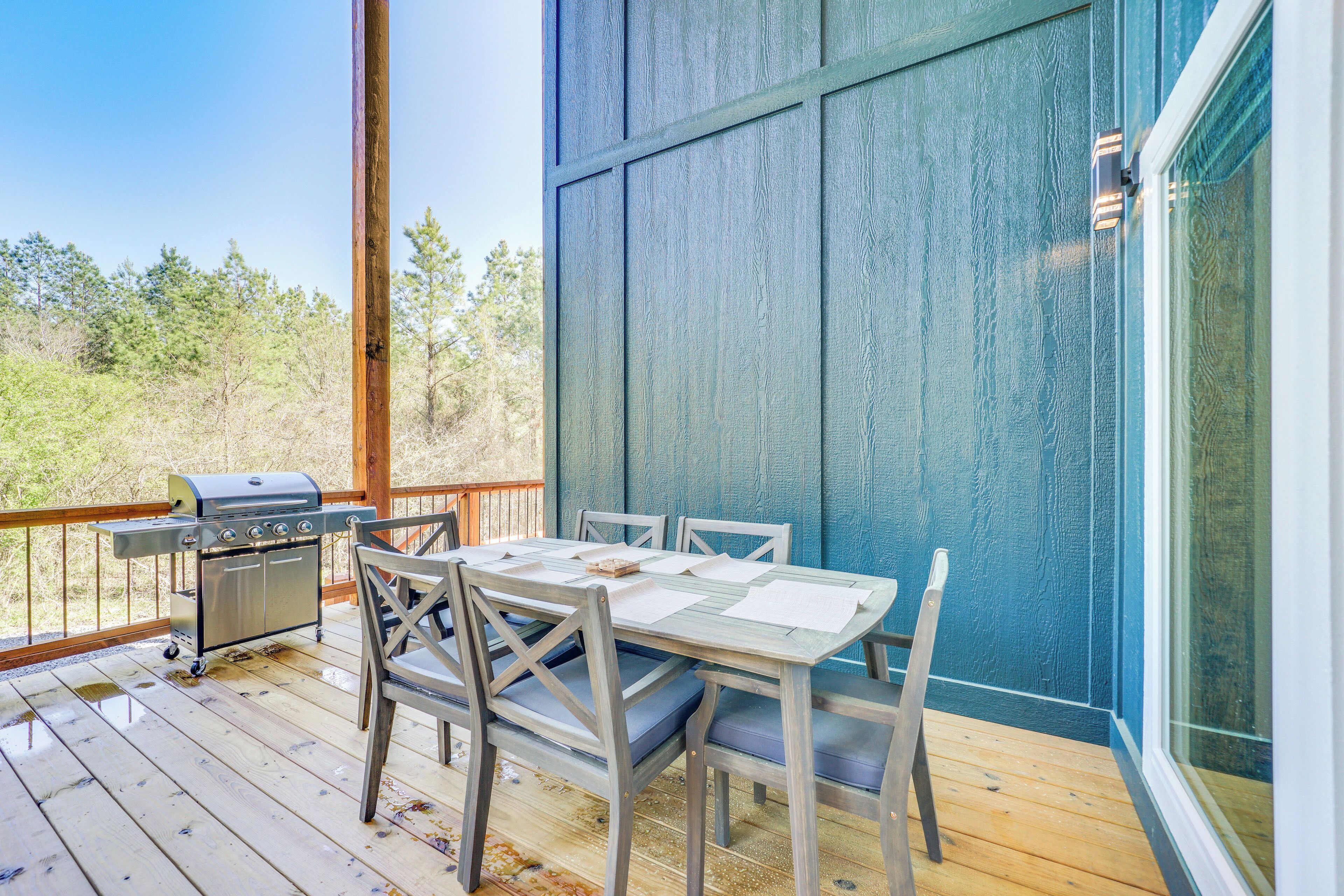 En udendørs terrasse med et træbord og stole, grill og udsigt til træer.