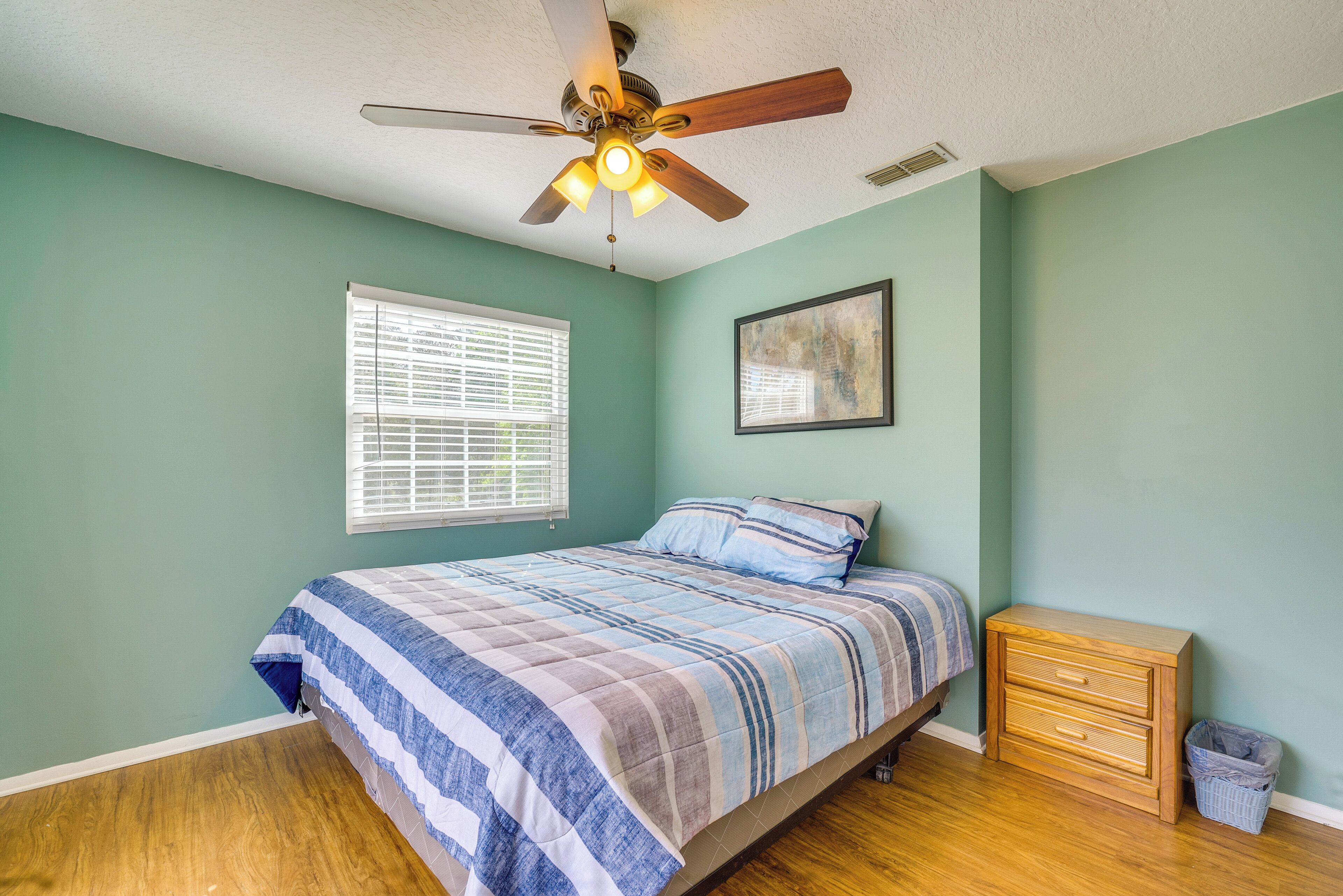 A bedroom with a bed, a ceiling fan, a nightstand, a window with blinds, and a framed picture on the wall.