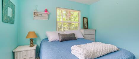 Un dormitorio con colcha azul, manta blanca, mesita de noche con lámpara y ventana con persianas.