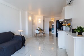 Ein modernes Wohnzimmer mit einer Couch, einem Esstisch mit Stühlen, einer Küchenzeile mit Kühlschrank und einer Topfpflanze.