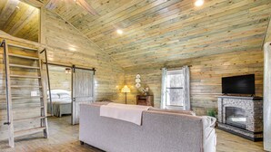 Interior acolhedor de uma cabana de madeira com sofá, lareira, televisão e escada para o mezanino.