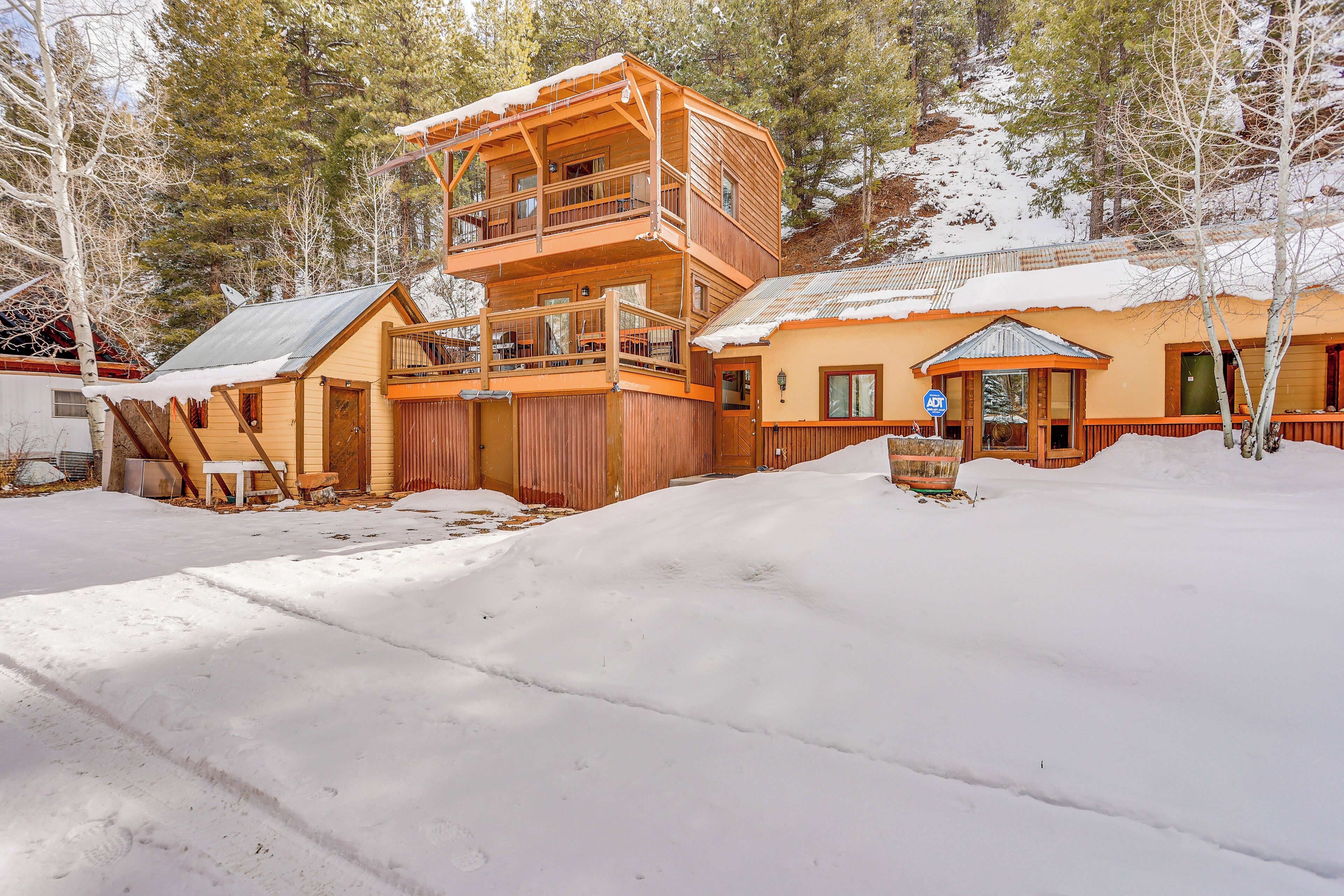 A two-story wooden cabin with a balcony, surrounded by snow-covered ground and trees.
