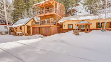 A two-story wooden cabin with a balcony, surrounded by snow-covered ground and trees.