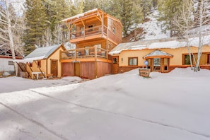 A two-story wooden cabin with a balcony, surrounded by snow-covered ground and trees.