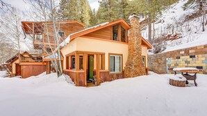 Una cabaña moderna de dos pisos con chimenea de piedra, terraza de madera y un paisaje cubierto de nieve.