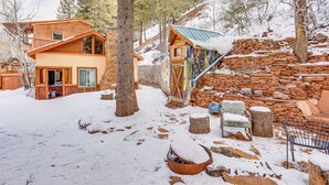 Una cabaña de madera con pared de piedra y techo metálico, rodeada de terreno y árboles cubiertos de nieve.