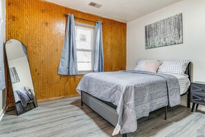 Een slaapkamer met een bed, houten paneelwanden, een raam met gordijnen, een spiegel en een kunstwerk aan de muur.