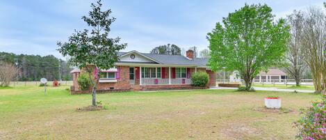 A single-story brick house with a porch, surrounded by a grassy yard and trees.